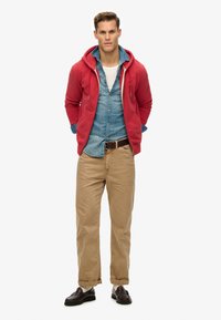 Felpa con zip rossa sopra una camicia in chambray azzurro chiaro, abbinata a pantaloni khaki e mocassini scuri. Design semplice, vestibilità casual, texture morbida.