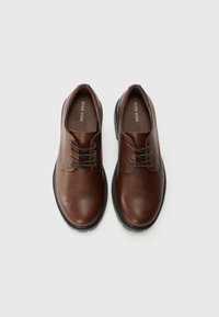 Zapatos de vestir de cuero marrón con textura suave, diseño de cordones y suela de goma negra contrastante, con una marca mínima en el interior.