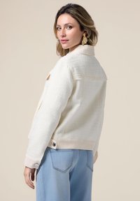 Giacca bianca texture con colletto e polsini di morbido crema, dotata di bottoni e dal design corto. Indossata con jeans azzurri chiari.
