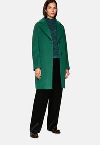 Grüner Wollmantel mit Reverskragen und zwei Knöpfen, getragen über einem blauen strukturierten Pullover, kombiniert mit schwarzen Hosen und Schuhen mit Tiermuster.