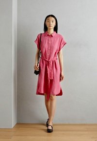 Abito camicia in lino rosa con maniche corte, chiusura a bottoni frontale, vita legata e lunghezza fino al ginocchio, abbinato a scarpe flat nere e una borsa a spalla.