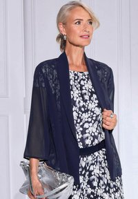 Marine doorzichtig vest over een bloemenprint jurk in donkerblauw en wit. Geaccessoreerd met een zilveren clutch en een statementring.