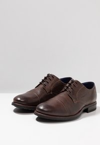 Paire de chaussures habillées en cuir marron à lacets, bout droit et coutures discrètes, présentée sur une surface blanche avec un fond uni.