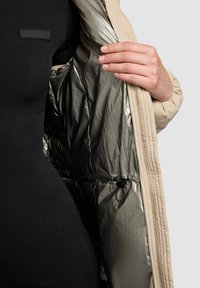 Veste extérieure beige avec une doublure argentée brillante, présentant un matériau texturé à l'intérieur et un cordon de serrage réglable à la taille.