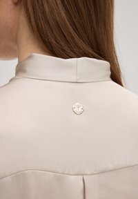 Lichtbeige blouse met een gladde textuur, voorzien van een kraag en een zilveren cirkelvormige logospeld aan de achterkant. Fijne plooidetail bij de zoom.