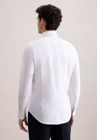 Man met krullend zwart haar, gekleed in een aansluitend wit overhemd met lange mouwen en donkere broeken, gezien van achteren tegen een effen achtergrond.