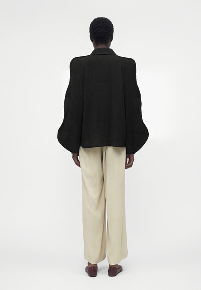 Άτομο που στέκεται γυρισμένο με την πλάτη φορώντας μαύρο υφασμάτινο μπουφάν με φαρδιά μανίκια, μπεζ παντελόνι φαρδιάς γραμμής και καφέ παπούτσια σε λευκό φόντο.