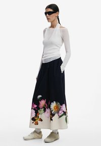 Lange marineblauwe rok met bloemenprint aan de zoom, gecombineerd met een aansluitend wit topje en beige sneakers. Het model draagt een donkere zonnebril.