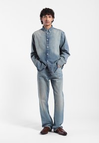 Denimowy zestaw składający się z jasnoniebieskiej koszuli zapinanej na guziki i dopasowanych dżinsów, obie z miękkiej tkaniny. Całość dopełniają brązowe buty.