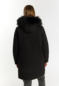 Sort parka-jakke med faux fur trim rundt hætten, med en ensfarvet design og en mellemlang længde, båret over grå bukser.