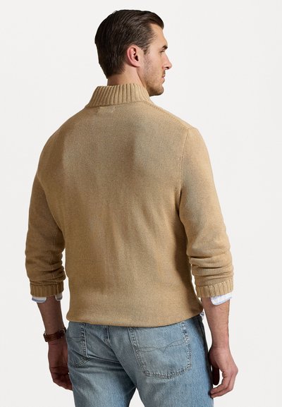 Beige stickad tröja med hög krage och ribbade ärmslut, med ett strukturerat mönster. Bärs över ljusblå denimjeans.