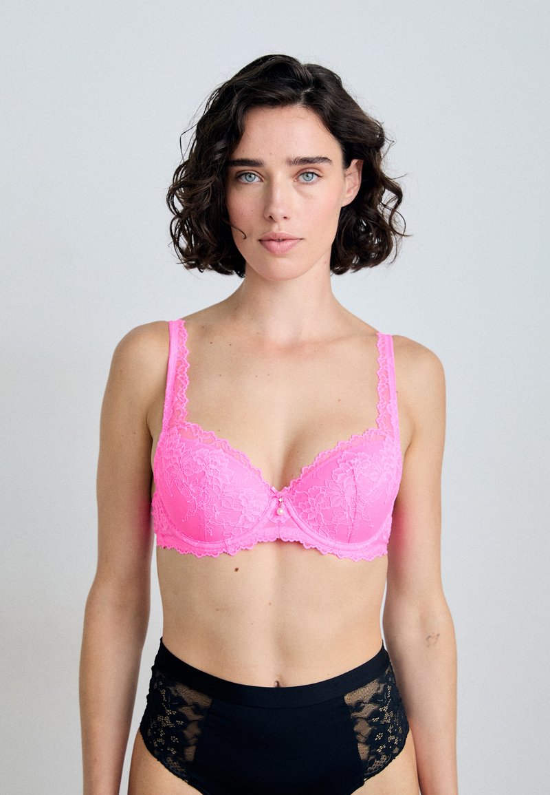 Rosa Spitzen-BH mit Unterdraht, floralem Muster, gewellten Kanten und einem zentralen dekorativen Element. Getragen mit schwarzen hochgeschnittenen Panties.