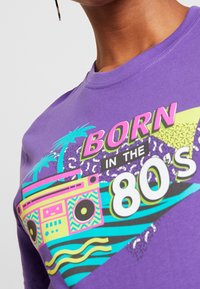 Lila t-shirt med grafisk tryck av en retro boombox, palmträd och texten "FÖDD PÅ 80-TALET" i livliga färger. Mjuk bomullstextur.