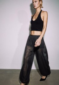 Crna majica na vezanje oko vrata s dubokim izrezom uparena s prozirnim crnim širokim hlačama s vertikalnim prugama i raspršenim sjajnim detaljima.