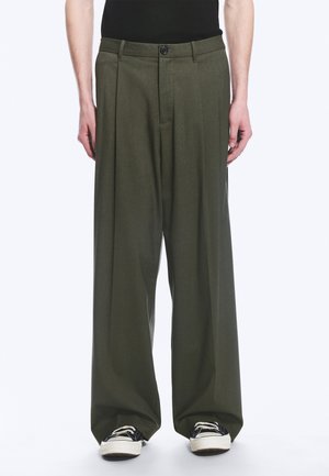 Homme portant un pantalon ample vert olive et des baskets noires à semelles blanches, debout devant un fond clair uni.