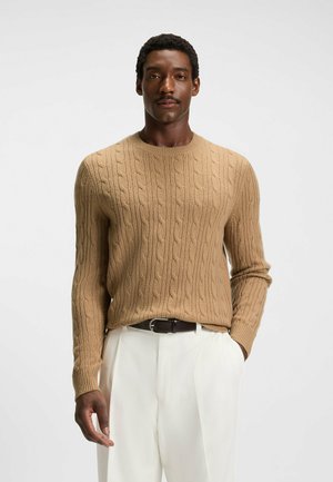 Maglione beige a maglia cable con scollatura rotonda, maniche lunghe e polsini a coste, abbinato a pantaloni bianchi sartoriali e una cintura marrone.