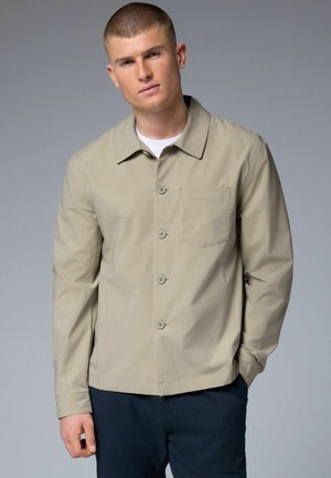 Beige jacket with a collar, front buttons, and a chest pocket. Features long sleeves and a smooth, matte texture. Worn over a white shirt.