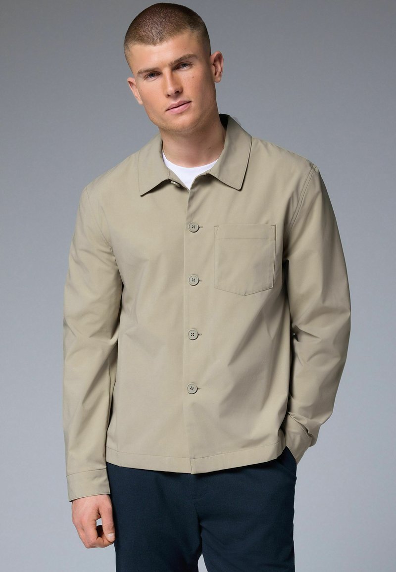 Beige jacket with a collar, front buttons, and a chest pocket. Features long sleeves and a smooth, matte texture. Worn over a white shirt.