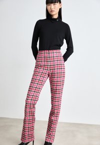 Un haut noir à col roulé associé à un pantalon taille haute à motif pied-de-poule rose, présentant une silhouette ajustée et de élégantes talons noirs.