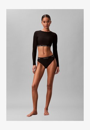 Vrouw staat op blote voeten, draagt een zwarte crop top met lange mouwen en bijpassend hoog uitgesneden ondergoed met zichtbare Calvin Klein-logo's.