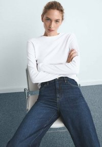 Vrouw met licht haar dat in een staart is gebonden, zit op een stoel met de armen over elkaar, gekleed in een witte lange mouwen top en donkerblauwe high-waisted jeans.