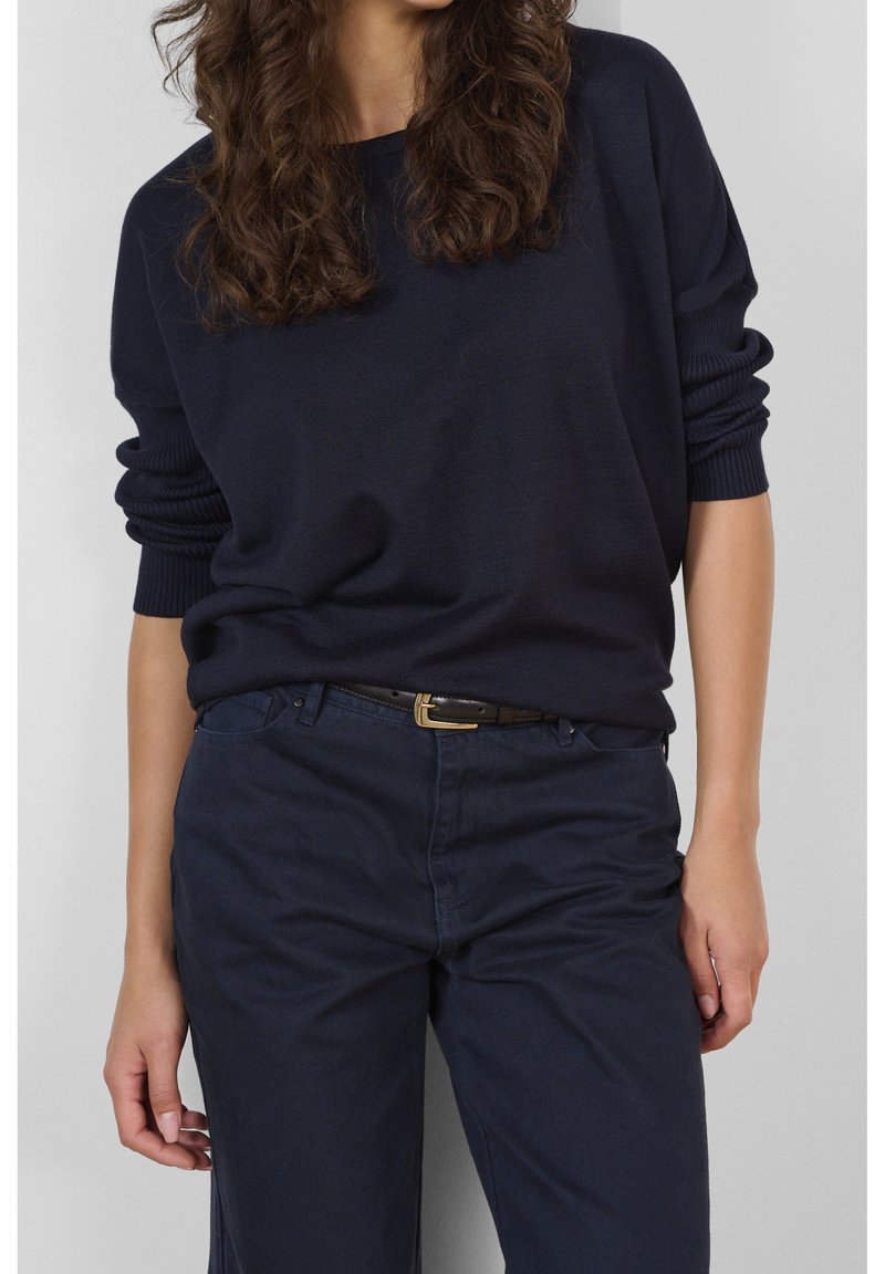 Femme portant un pull bleu marine foncé aux manches retroussées, rentré dans un pantalon bleu marine foncé avec une ceinture noire, debout sur un fond gris.