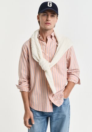 Junger Mann mit marineblauer Kappe mit G, rosa-blau gestreiftem Hemd mit Knöpfen, weißem Pullover über den Schultern und blauen Jeans.