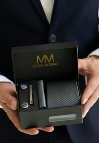 Graues Accessoire-Set in einer schwarzen Box, bestehend aus gestreifter Krawatte, Manschettenknöpfen und Krawattenschieber. Goldenes Logo "MASSI MORINO" auf dem Deckel der Box.