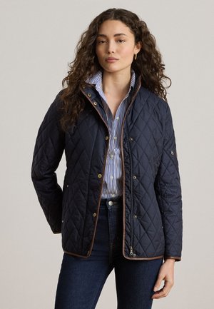 Femme aux longs cheveux bouclés portant une veste matelassée bleu marine par-dessus une chemise rayée bleue et blanche et un jean foncé, debout devant un fond uni.