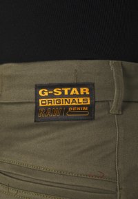 Olivgröna byxor med G-Star Originals svart etikett som har gul och orange text. Stickad detaljer synlig på tyget.