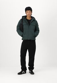 Dark green padded jacket with a zip front, two chest pockets, and a hood, paired with black pants and black sneakers with white stripes.