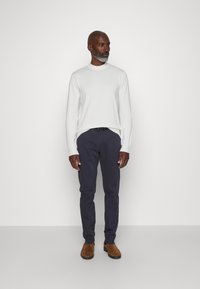 Baltas megztinis, tamsiai mėlyni kelnes, rudi zomšiniai batai. Megztinis turi apvalų kaklą; kelnes tiesaus kirpimo. Paprastas ir švarus dizainas.