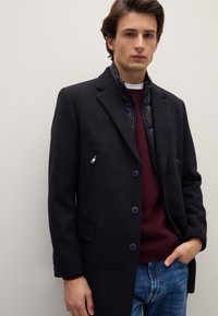 Cappotto lungo in lana scura con colletto a rever, bottoni monopetto e tasca laterale con zip, indossato sopra un maglione a maglia bordeaux.