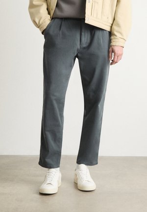Grey cotton trousers with a relaxed fit and pleats, paired with white sneakers. The background is neutral and minimalistic.