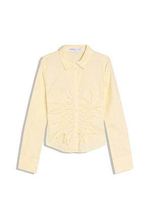 Vaaleankeltainen, pitkähihainen kauluksellinen paitapusero, jossa on napit edessä ja ryppyiset etuosan yksityiskohdat, jotka on sidottu pienillä ruseteilla vyötäröllä.