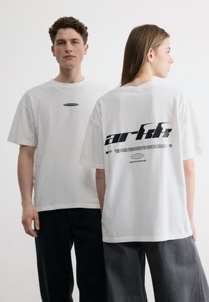 Due modelle che indossano magliette bianche oversize, una rivolta verso il davanti con il logo sul petto, l'altra che mostra un grande testo "arkk" sulla schiena.