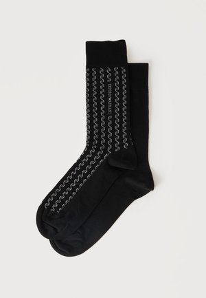 Paire de chaussettes noires mi-mollet, dont une présente des bandes réfléchissantes verticales en zigzag et le texte "Emporio Armani" sur un fond uni clair.