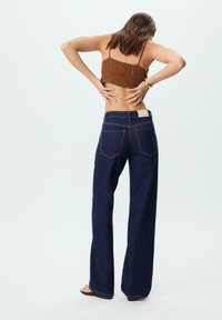 Jean à taille haute, bleu foncé, avec des jambes larges en denim, comprenant deux poches arrière et des coutures visibles. Modèle vu de dos, portant un haut marron.