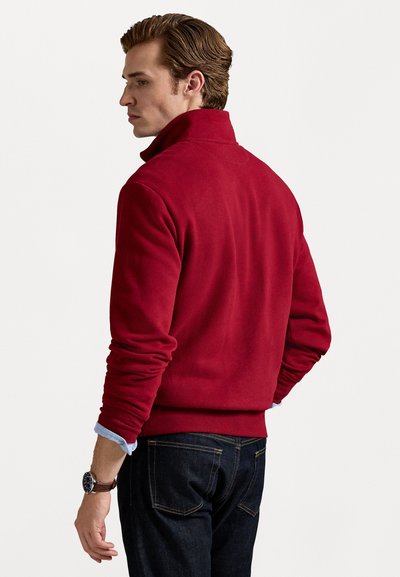 Rdeča flis majica z visokim ovratnikom, rebrastimi manšetami in robom, v kombinaciji s temnimi kavbojkami. Tkanina se zdi mehka in teksturirana.