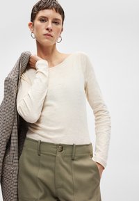 Kremo spalvos ilgomis rankovėmis palaidinė su apvaliu kaklu, derinama su alyvuogių žalumos kelnėmis. Ant vieno peties užmestas margas švarkas.