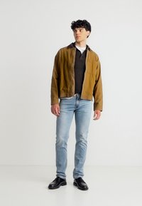 Νέος άνδρας στέκεται κοιτάζοντας δεξιά, φοράει καφέ μπουφάν, μαύρο πουκάμισο, ανοιχτό μπλε τζιν και μαύρα δερμάτινα παπούτσια μπροστά σε απλό λευκό φόντο.