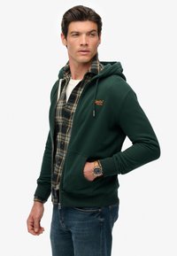 Hombre con sudadera de cremallera verde oscuro sobre camisa de cuadros y jeans, con un reloj de pulsera de cuero en la muñeca izquierda, mirando hacia un lado.