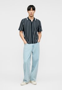 Νέος άνδρας με μαύρα ίσια μαλλιά, φορούσε μπλε σκούρο κοντομάνικο πουκάμισο με λευκές κάθετες ρίγες, ανοιχτό μπλε τζιν παντελόνι φαρδύ και λευκά αθλητικά παπούτσια.