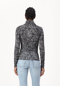 Langsleeve top gemaakt van doorzichtig zwart materiaal met een wit abstract patroon. Heeft een hoge kraag en plooien aan de zijkanten. Draagt bij lichte blauwe jeans.