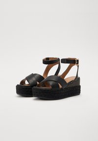 Schwarze Plattform-Sandalen mit Kreuzband-Design, aus Leder gefertigt. Mit Knöchelriemen und einer gewobenen, korkähnlichen Sohle.