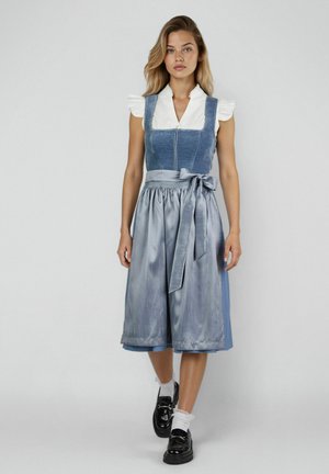 Vrouw staand, gekleed in een blauwe en zilveren traditionele jurk met een strikceintuur, wit geruffeld overhemd, witte sokken en zwarte loafers tegen een effen achtergrond.