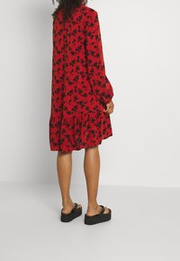 Vestido rojo con estampado floral negro, de mangas largas, con dobladillo en capas, hecho de tejido ligero. Combinado con sandalias de plataforma negras.