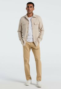 Beige knoopshirt met twee borstzakken, gedragen over een wit T-shirt, gecombineerd met lichtbruine broek en witte sneakers. Eenvoudige, geborstelde stof.