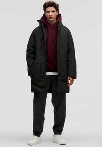Zwarte gewatteerde parka met meerdere zakken, gedragen over een donkerrode hoodie. De outfit heeft zwarte broek en witte sneakers.