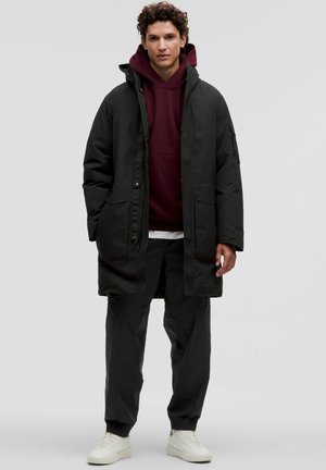 Sort polstret parka med flere lommer, båret over en mørkerød hættebluse. Outfittet har sorte bukser og hvide sneakers.
